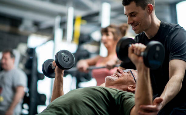 A senior man and a male trainer are indoors in a fitness center. They are wearing casual athletic clothing. The man is lifting weights with the guidance of the trainer.