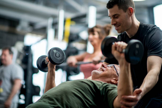A senior man and a male trainer are indoors in a fitness center. They are wearing casual athletic clothing. The man is lifting weights with the guidance of the trainer.